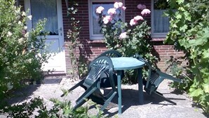 Una casa de ladrillo con techo de paja, una ventana adornada con rosas rosadas, y un pequeño espacio al aire libre con una mesa y dos sillas.