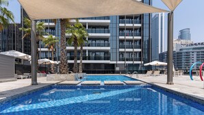A swimming pool with a tiled floor, surrounded by lounge chairs and umbrellas, with a modern building in the background. - Dubaiashome - Luxury Special Stay (Dubai)