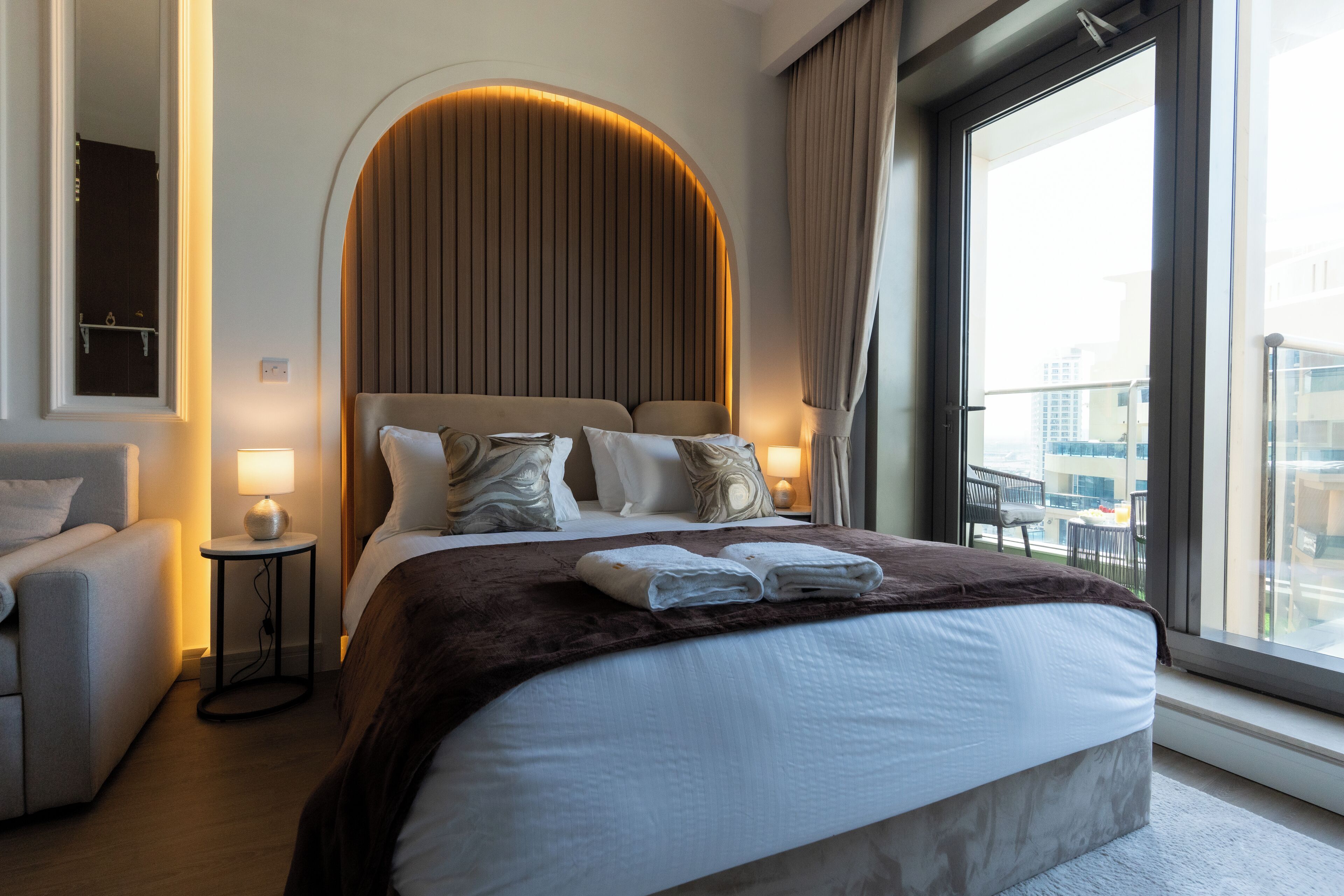 Habitación de hotel con una cama grande, un sofá, una mesita con lámpara y vistas a la ciudad a través de un ventanal amplio.