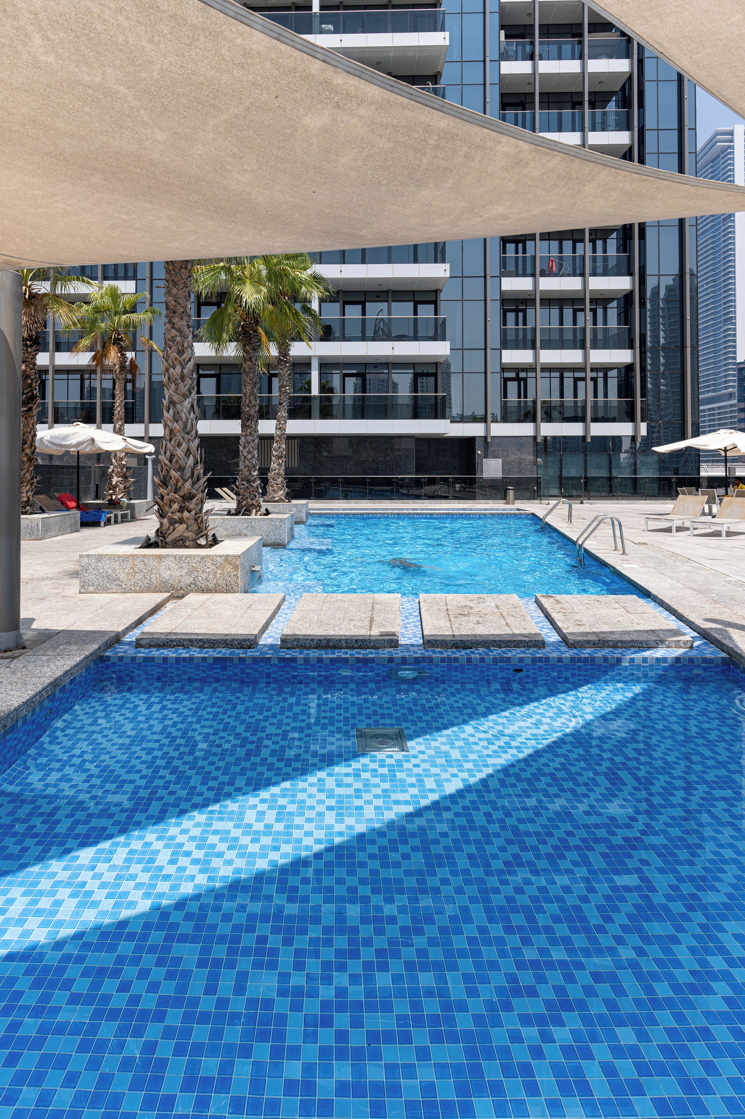 A swimming pool with clear blue tiles, surrounded by palm trees and lounge chairs, with a modern building in the background.