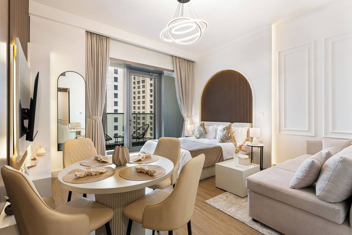 A modern hotel room with a bed, a sofa, a dining table with chairs, a TV, and a large window with curtains.