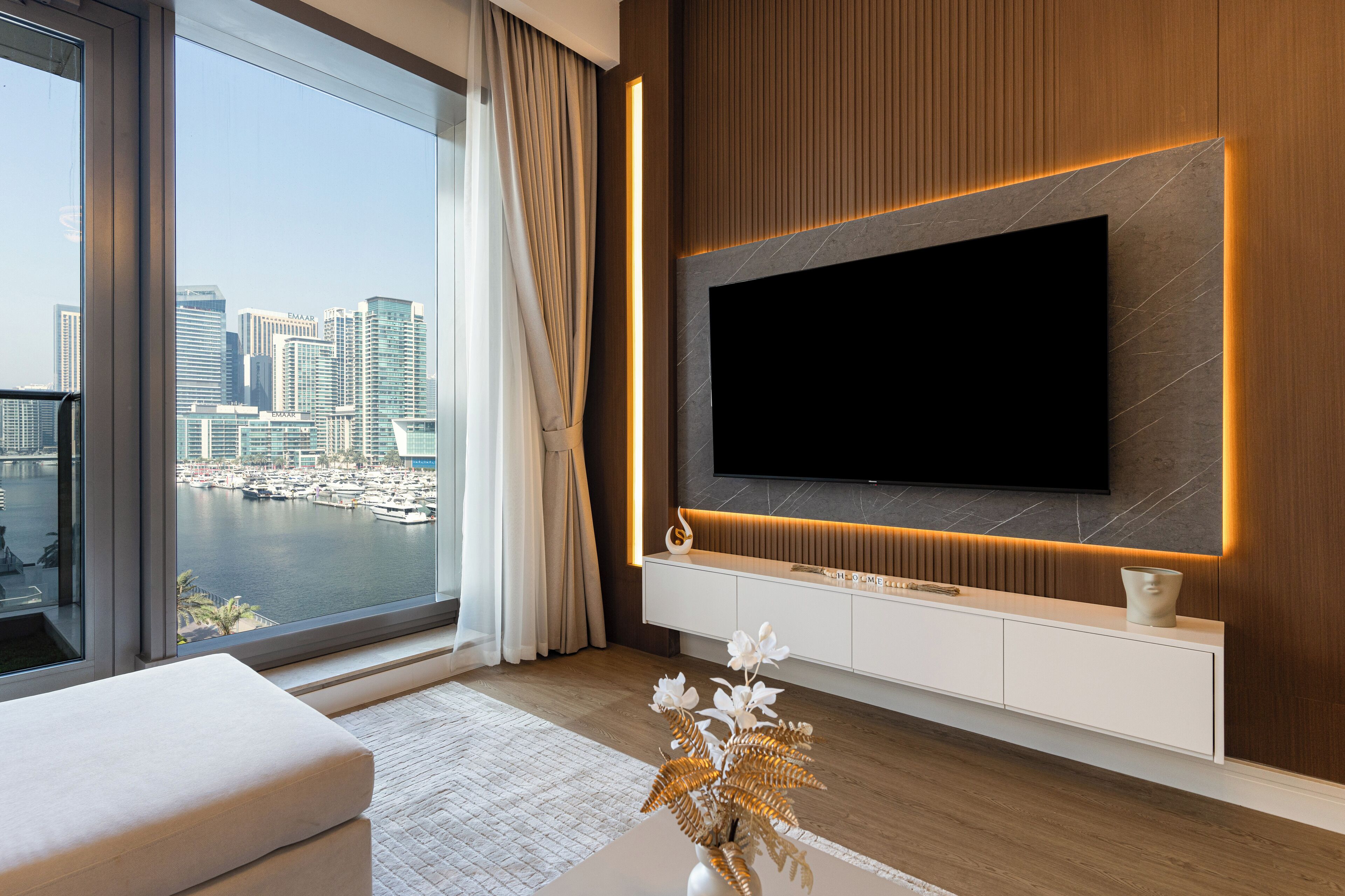 Un salon moderne doté d’un grand téléviseur, d’une console blanche et offrant une vue sur une marina et le paysage urbain.