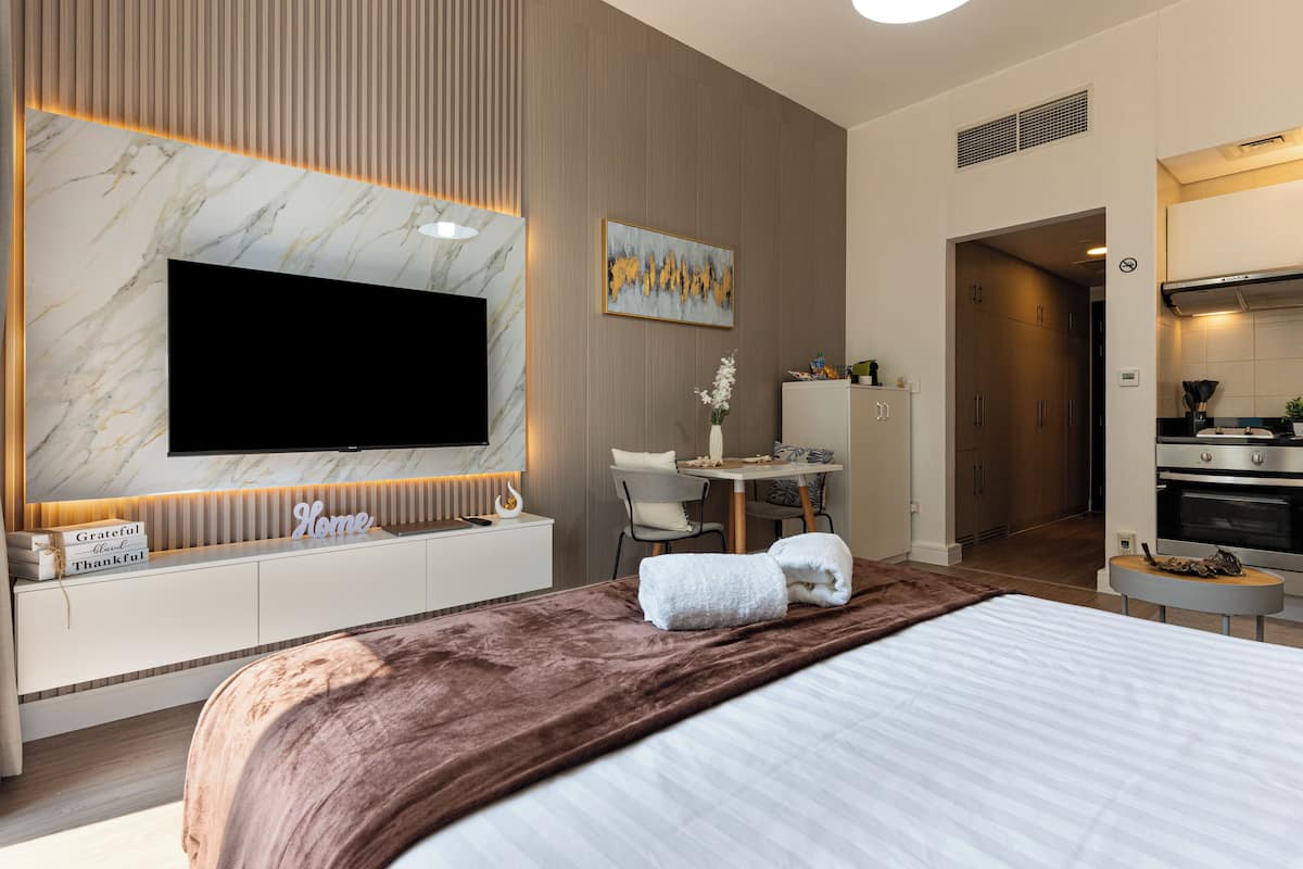 A modern hotel room with a large flat-screen TV, a marble accent wall, a dining area with a table and chairs, and a kitchenette.