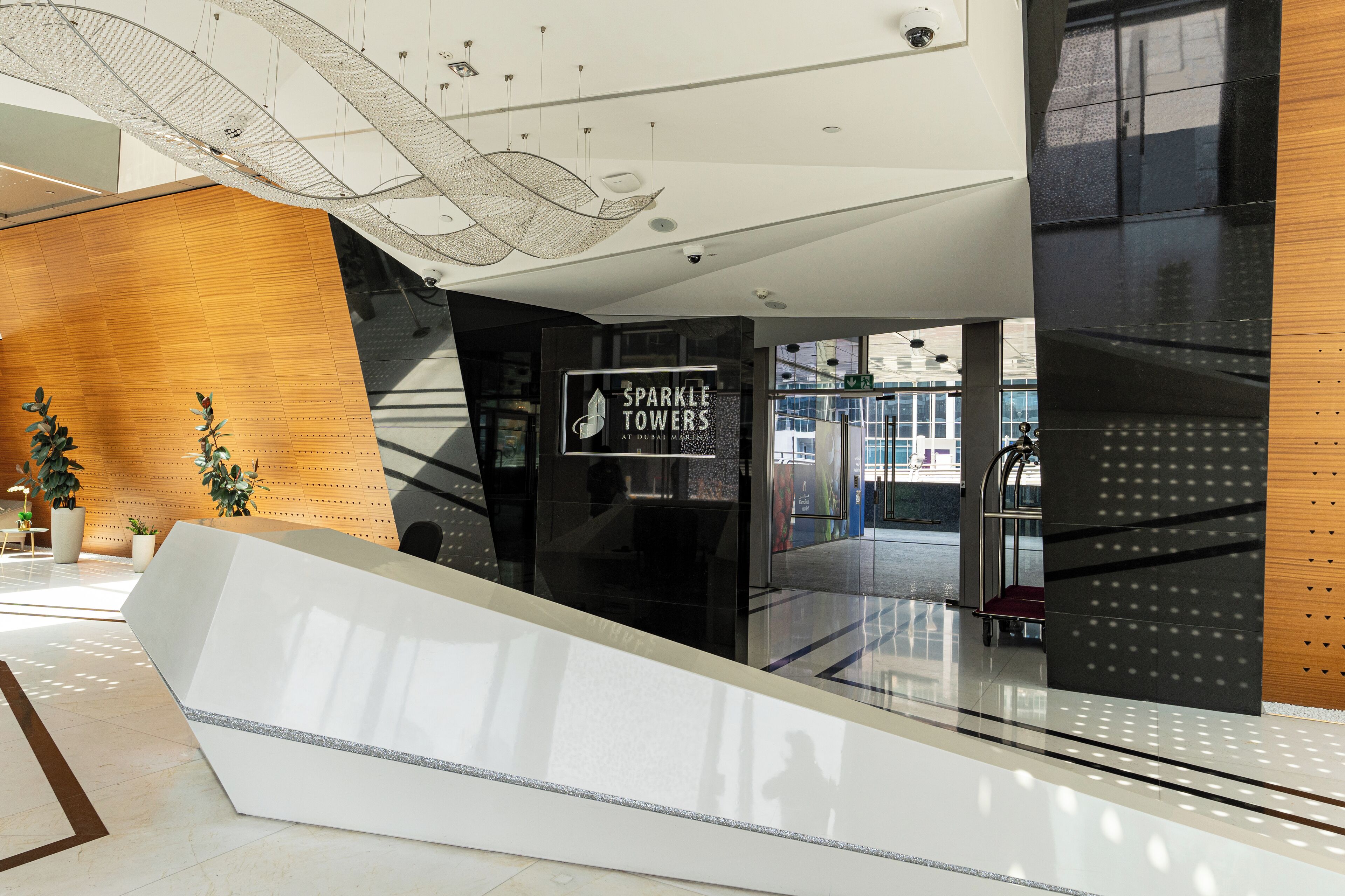 Una hall d'albergo moderna con una reception in legno, un ascensore in vetro e un cartello che indica 'Sparkle Towers at Dubai Marina'.