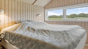 Ein Schlafzimmer mit Holzbalkendecke, einem großen Bett mit gemustertem Überwurf, einer Nachttischlampe und einem Fenster mit Blick auf Grünflächen.