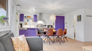 Een moderne woonkamer met een grijze bank, een eetgedeelte met paarse kasten, en een keuken met witte en paarse details.