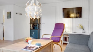 Een moderne woonkamer met een houten salontafel, een paarse fauteuil, een grijze bank, een witte, aan de muur bevestigde televisie en een bijzondere, sferische lamp.