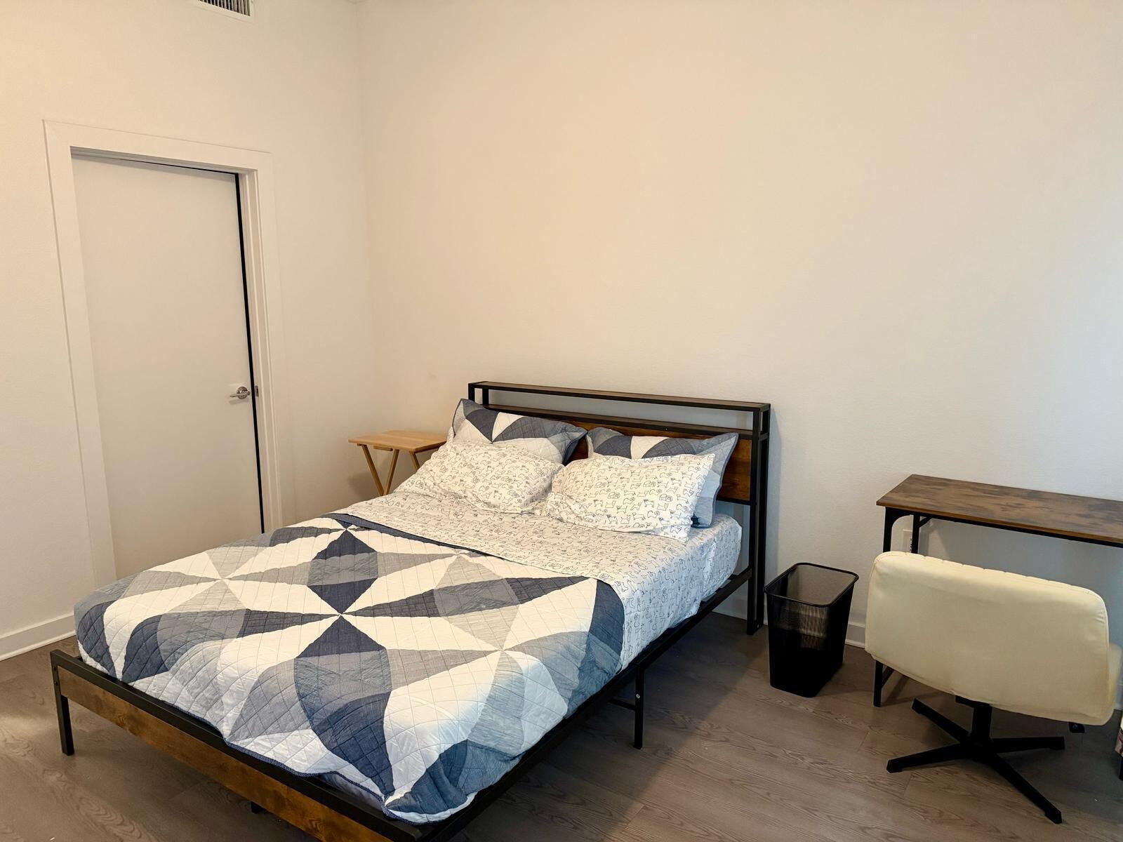 A bedroom with a bed, a small table, a chair, and a trash bin.