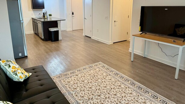 Een moderne woonkamer met een bank, een televisie op een dressoir, een keukenhoek met een koelkast, en een kamerbreed tapijt met een patroon.