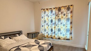 Een slaapkamer met een bed, een stoel, een kleine tafel en een raam met bloemen gordijnen.