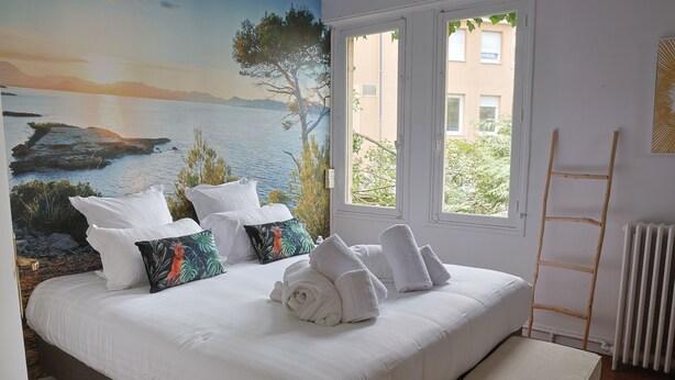 A bed with white linens, a bench, a mural of a coastal scene, and a window with a view of buildings.