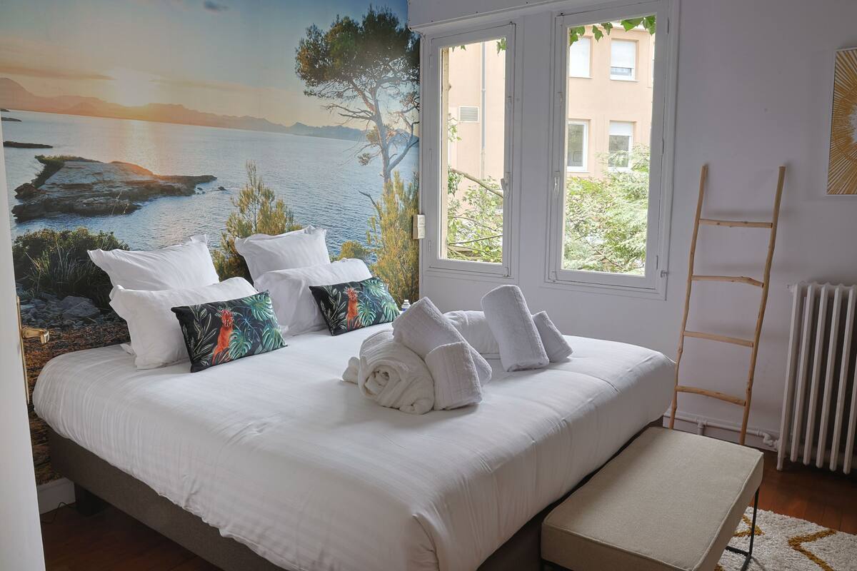 Een bed met witte lakens, een bankje, een muurschildering van een kustlandschap en een raam met uitzicht op gebouwen.