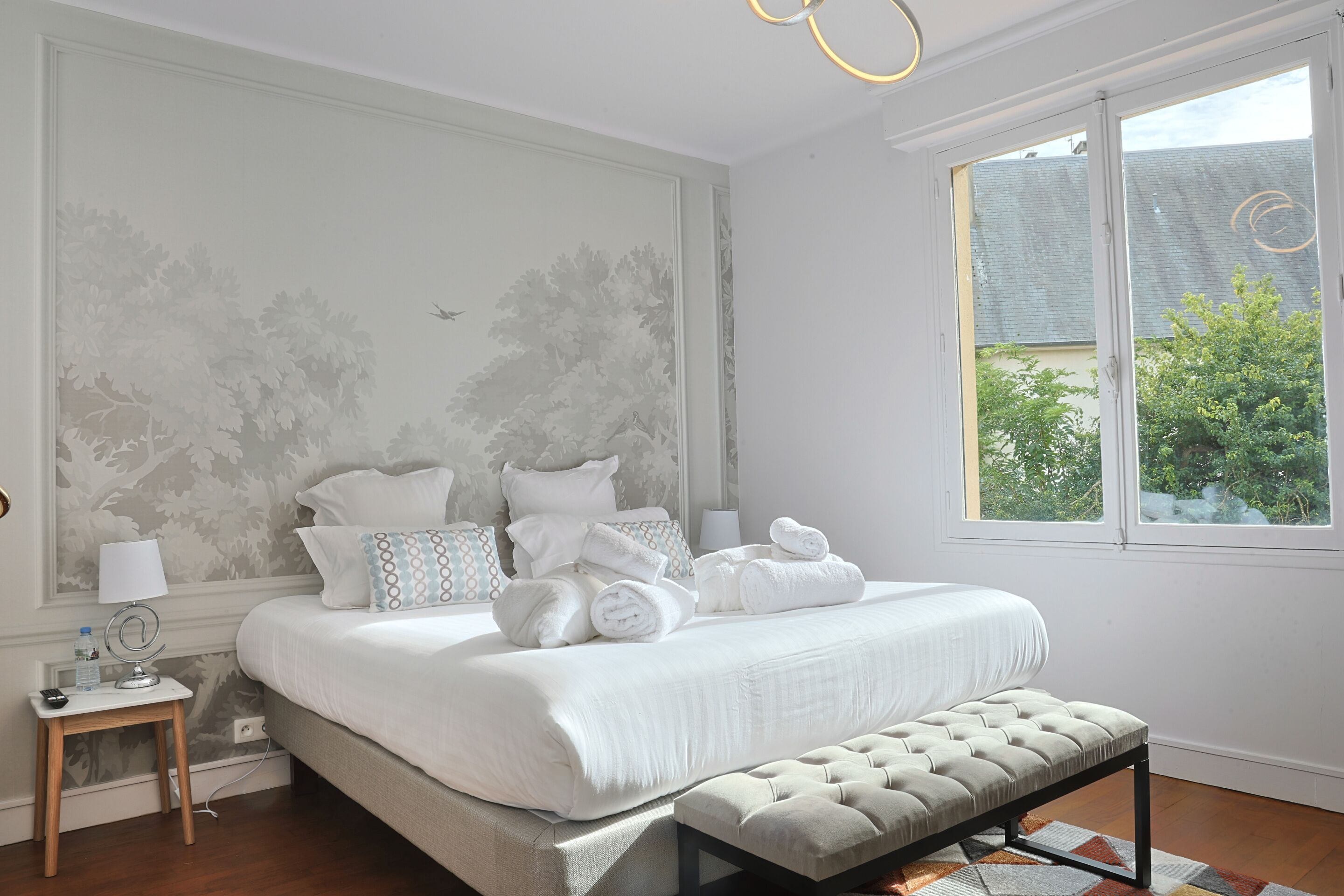 A bedroom with a large bed, bedside tables, a bench, and a window with a view of greenery.