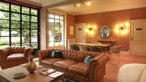 A spacious living area with a leather sofa, dining table, and chairs, large windows, and a wooden wall.