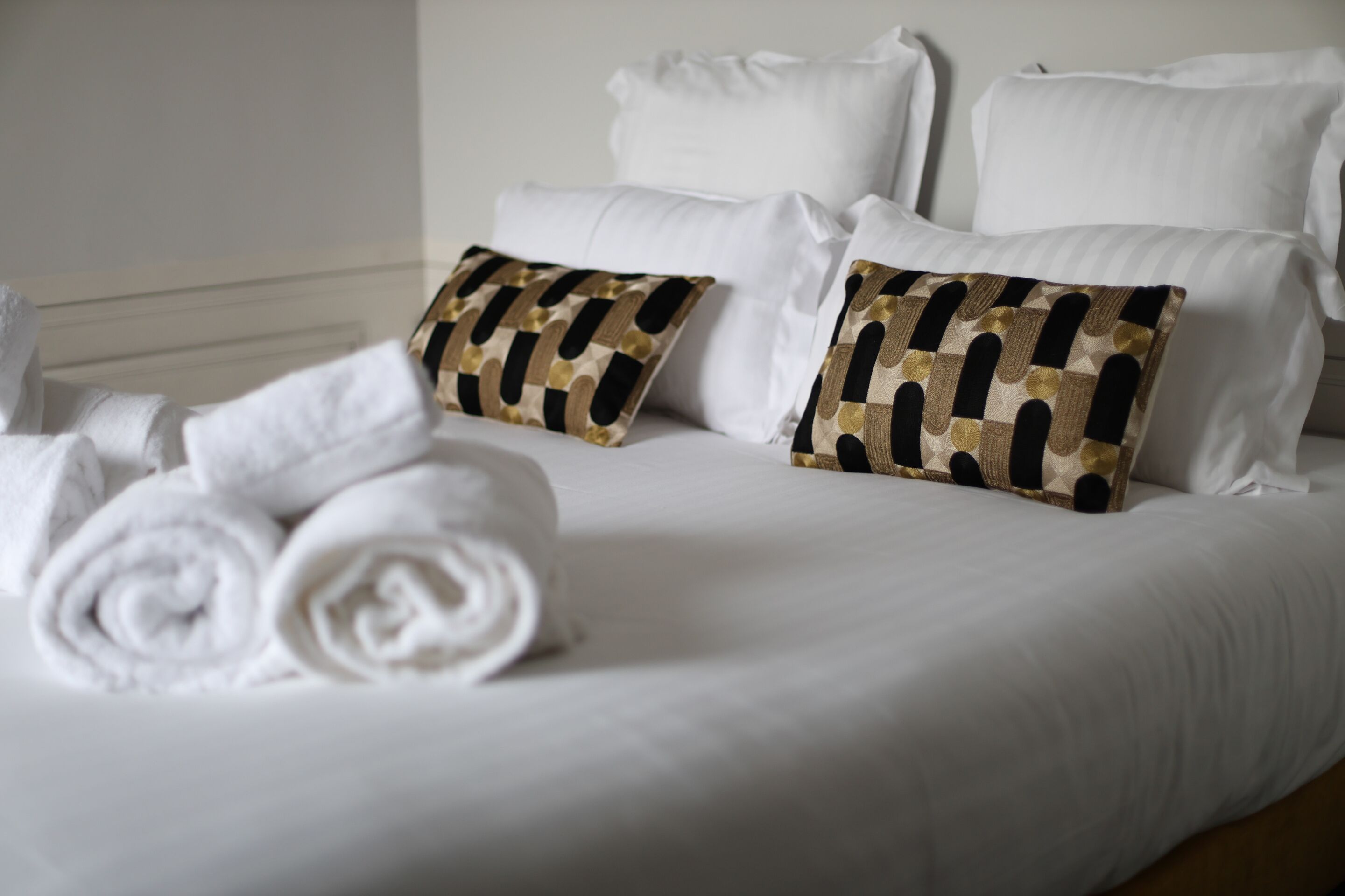 Una cama bien tendida con sábanas blancas, dos almohadas estampadas y toallas enrolladas.