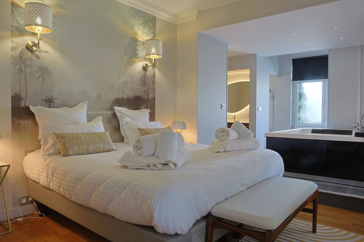 Een slaapkamer met een groot bed, nachtlampjes, een bankje, een badkamer met een badkuip en een spiegel.