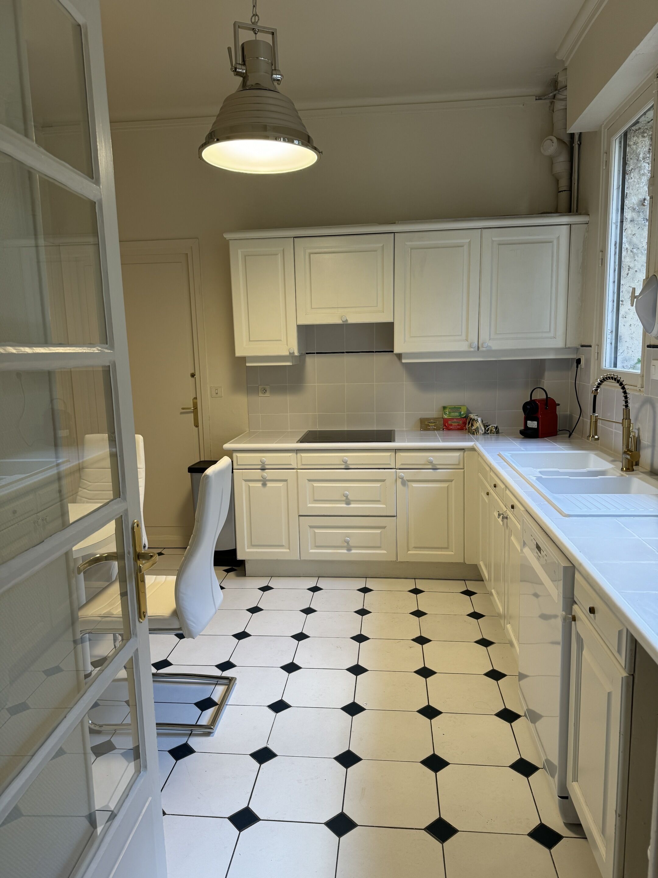 Una cocina con armarios blancos, suelo de baldosas blanco y negro y una lámpara colgante.