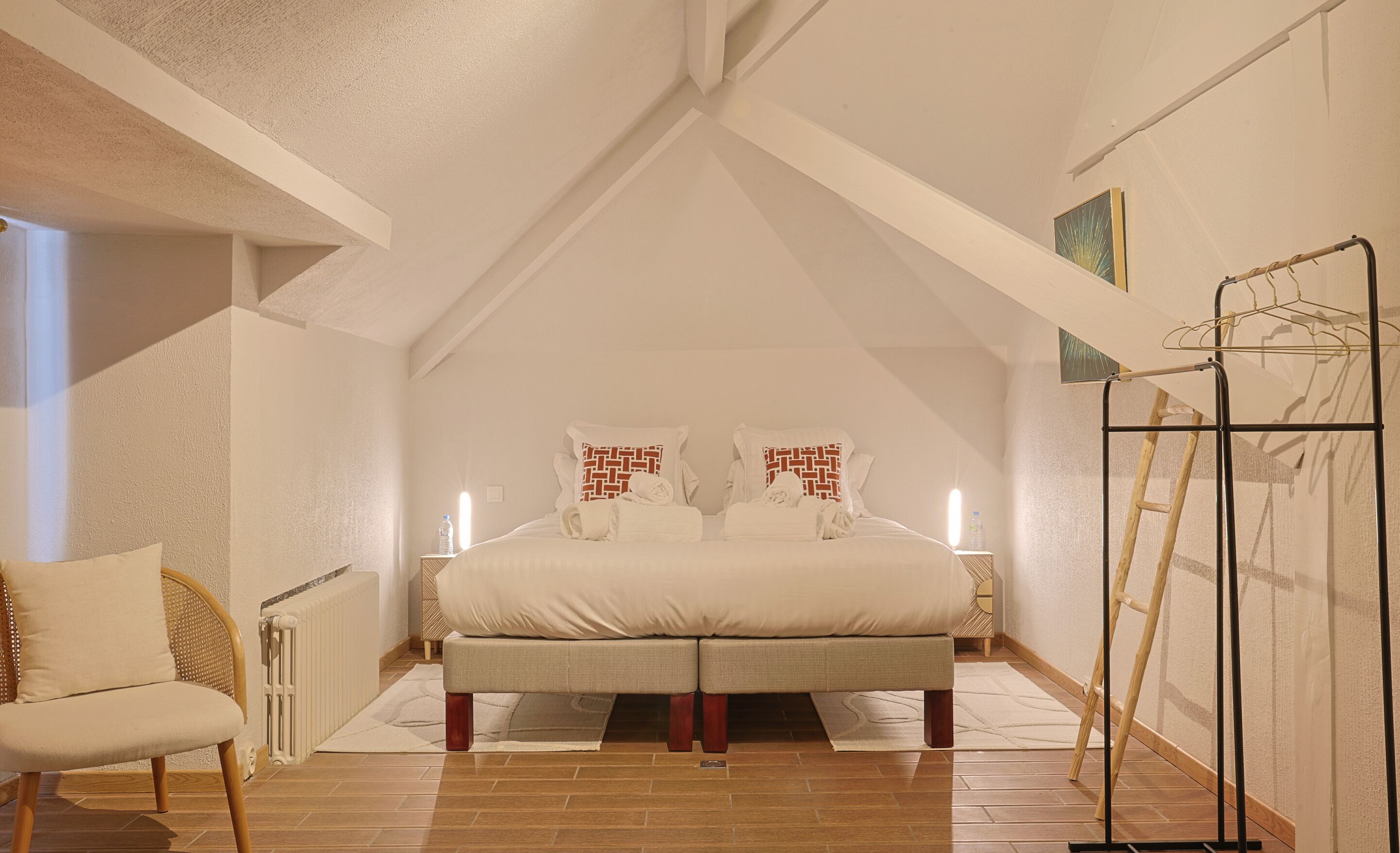 Une chambre à coucher avec un lit, deux tables de chevet, une chaise, un radiateur, une bouteille d’eau et une échelle.