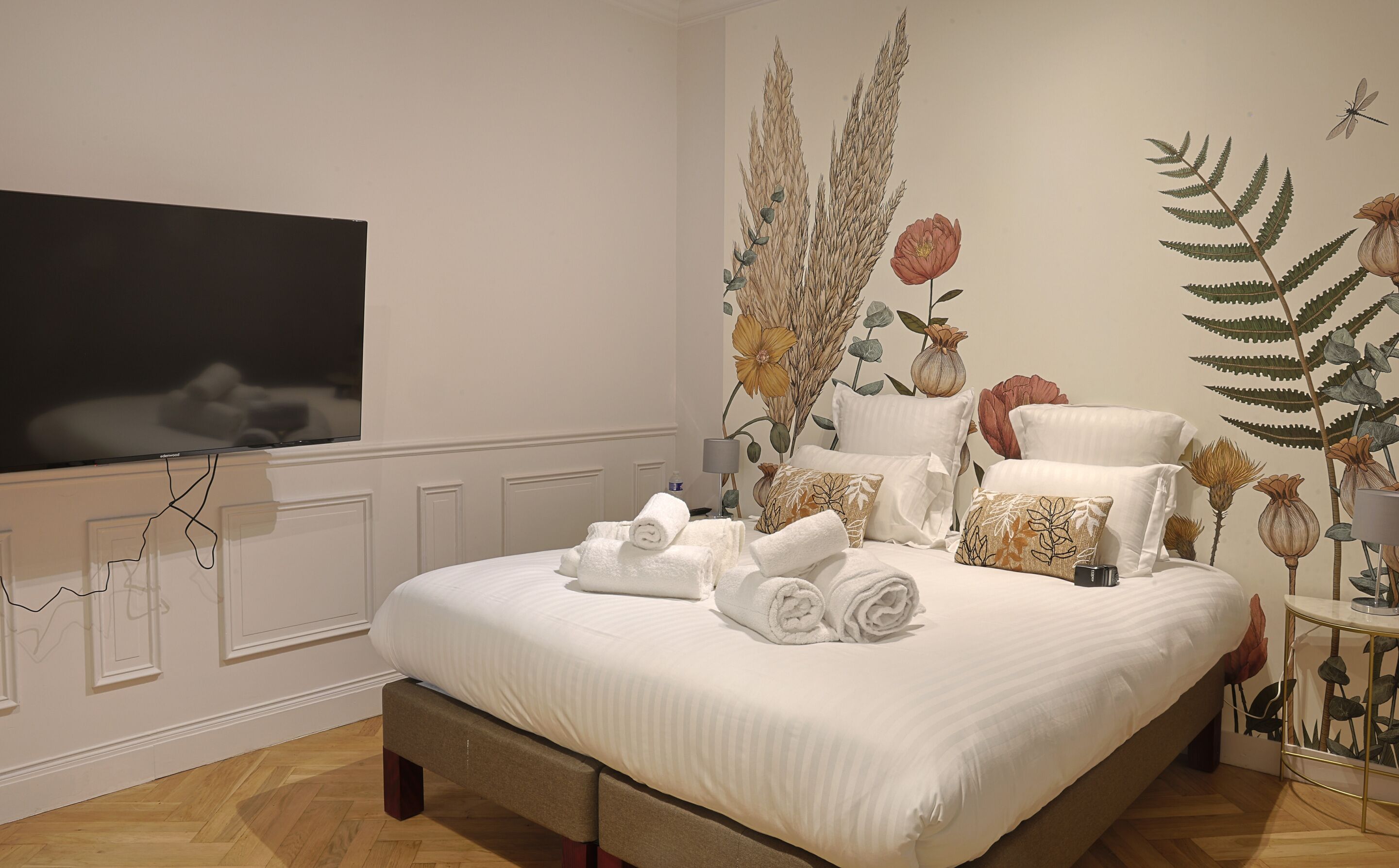 A neatly made bed with rolled towels, a flat-screen TV mounted on the wall, and a decorative wall mural with botanical elements.