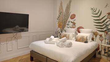 A neatly made bed with rolled towels, a flat-screen TV mounted on the wall, and a decorative wall mural with botanical elements.