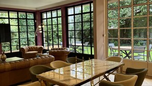 A spacious room with a large dining table, comfortable seating, and large windows offering a view of greenery.