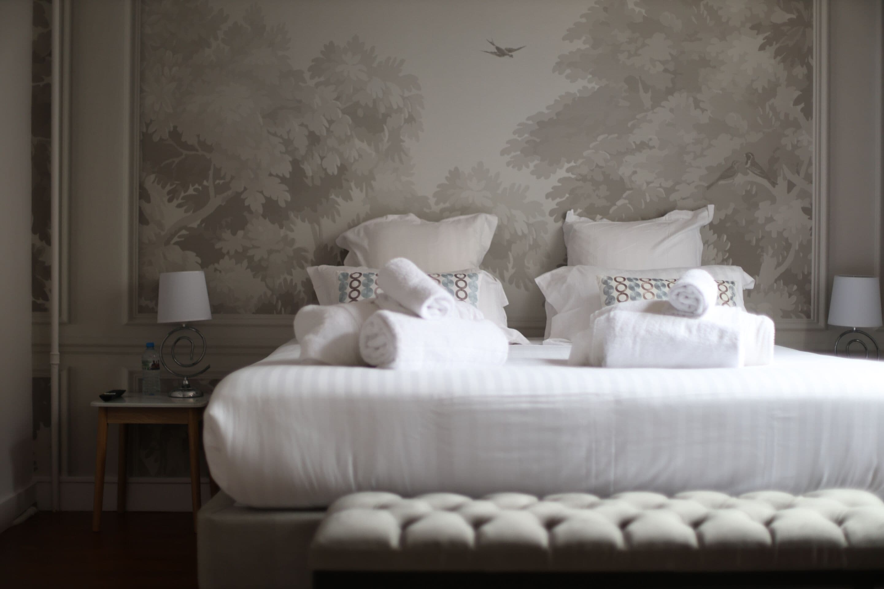 A neatly made bed with white linens and pillows, a small bedside table with a lamp, and a wall with a tree pattern.