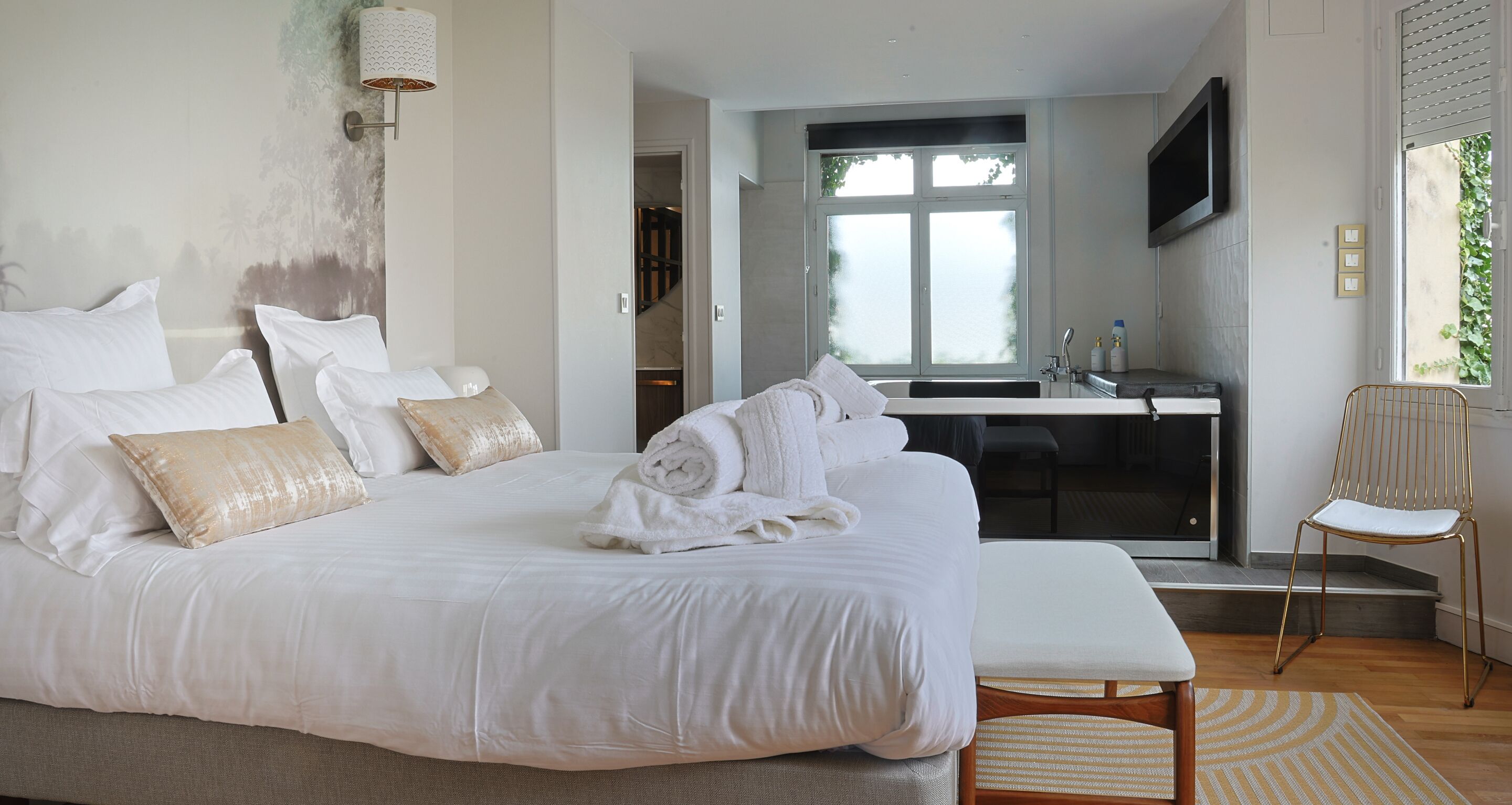 Quarto de hotel com uma cama grande, área de estar, banheiro com espelho e televisão fixada na parede.