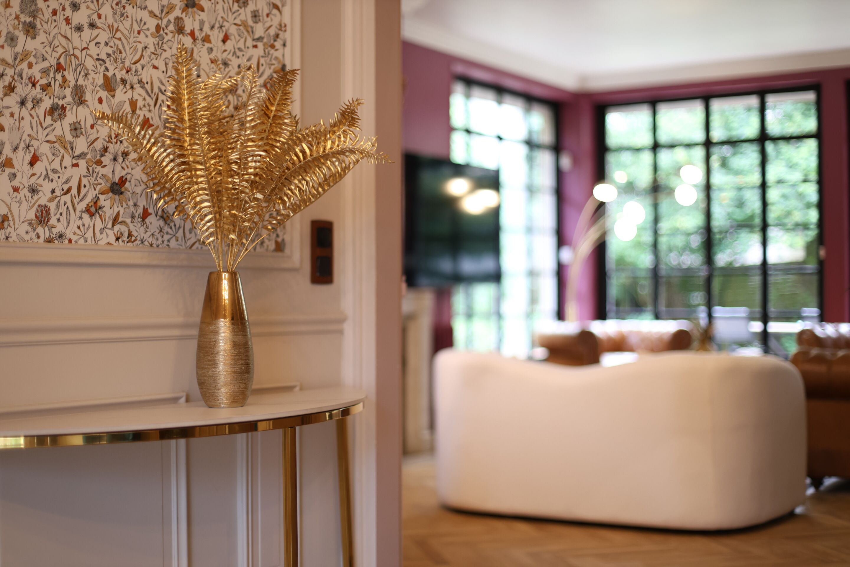 Una habitación con un sofá blanco, un jarrón dorado con una planta decorativa y un papel tapiz estampado.