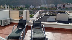 Una terraza en la azotea con dos sillones de descanso, una fuente de agua y vista a un paisaje urbano con edificios y una montaña al fondo.