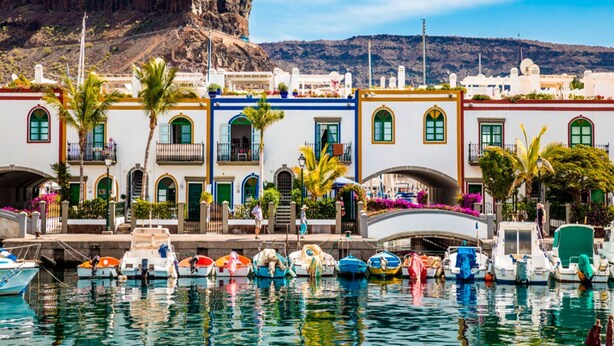 Un pueblo costero con edificios coloridos, un puerto deportivo con barcos y una montaña al fondo.