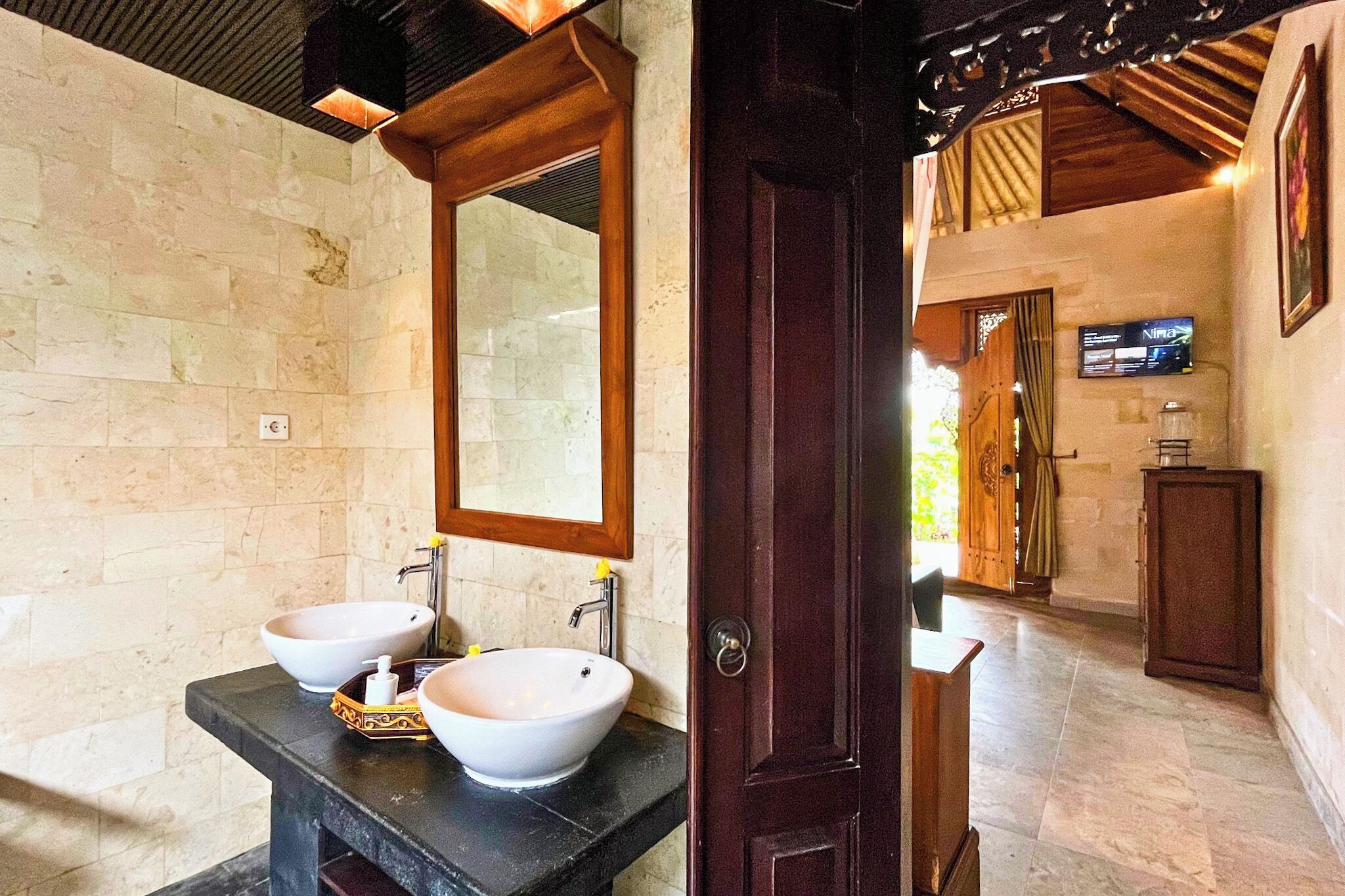 Un baño con dos lavamanos tipo isla blancos, un espejo grande y una puerta de madera que da a otra habitación.