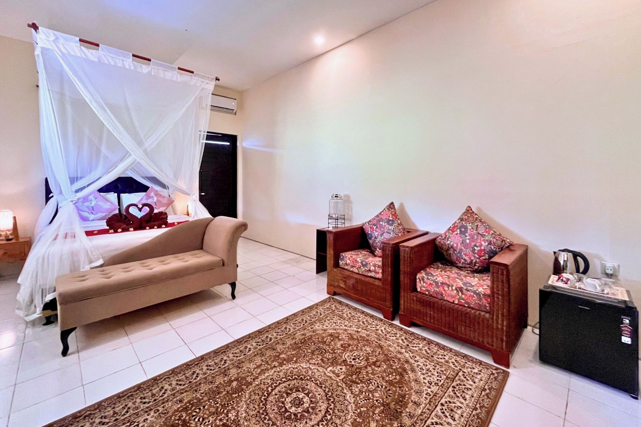 A room with a bed, a sofa, two armchairs, a small table, a decorative rug, and a minibar.