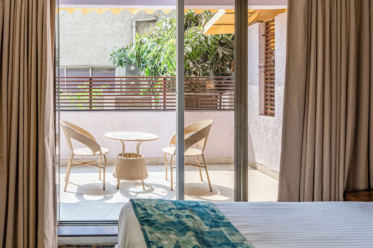 A hotel room with a bed, a view of an outdoor balcony with wicker chairs and a table, and a window with curtains.