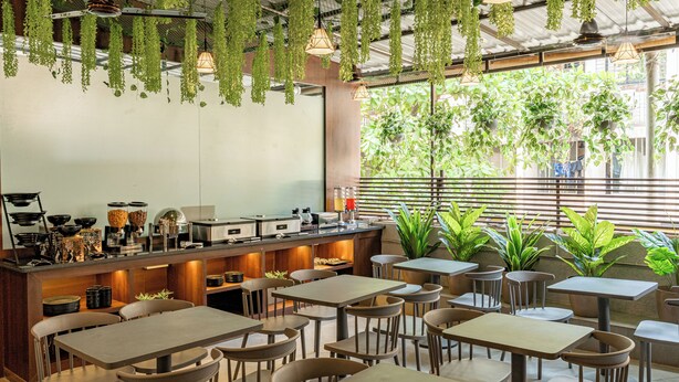 A restaurant with multiple tables and chairs, a counter with kitchen equipment, hanging plants, and large windows with views of greenery. - Hotel Dakshin Grandeur (Navi Mumbai)