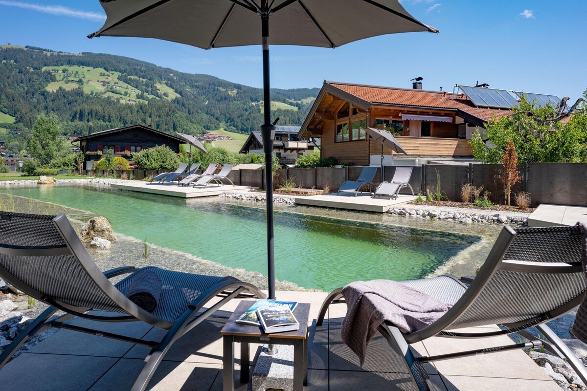 A poolside area with lounge chairs, an umbrella, and a view of a mountain landscape.
