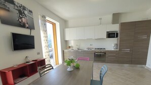 Una cocina moderna con armarios blancos, un microondas y una estufa. Hay una mesa de comedor con sillas, un televisor montado en la pared y un estante rojo con una lámpara.