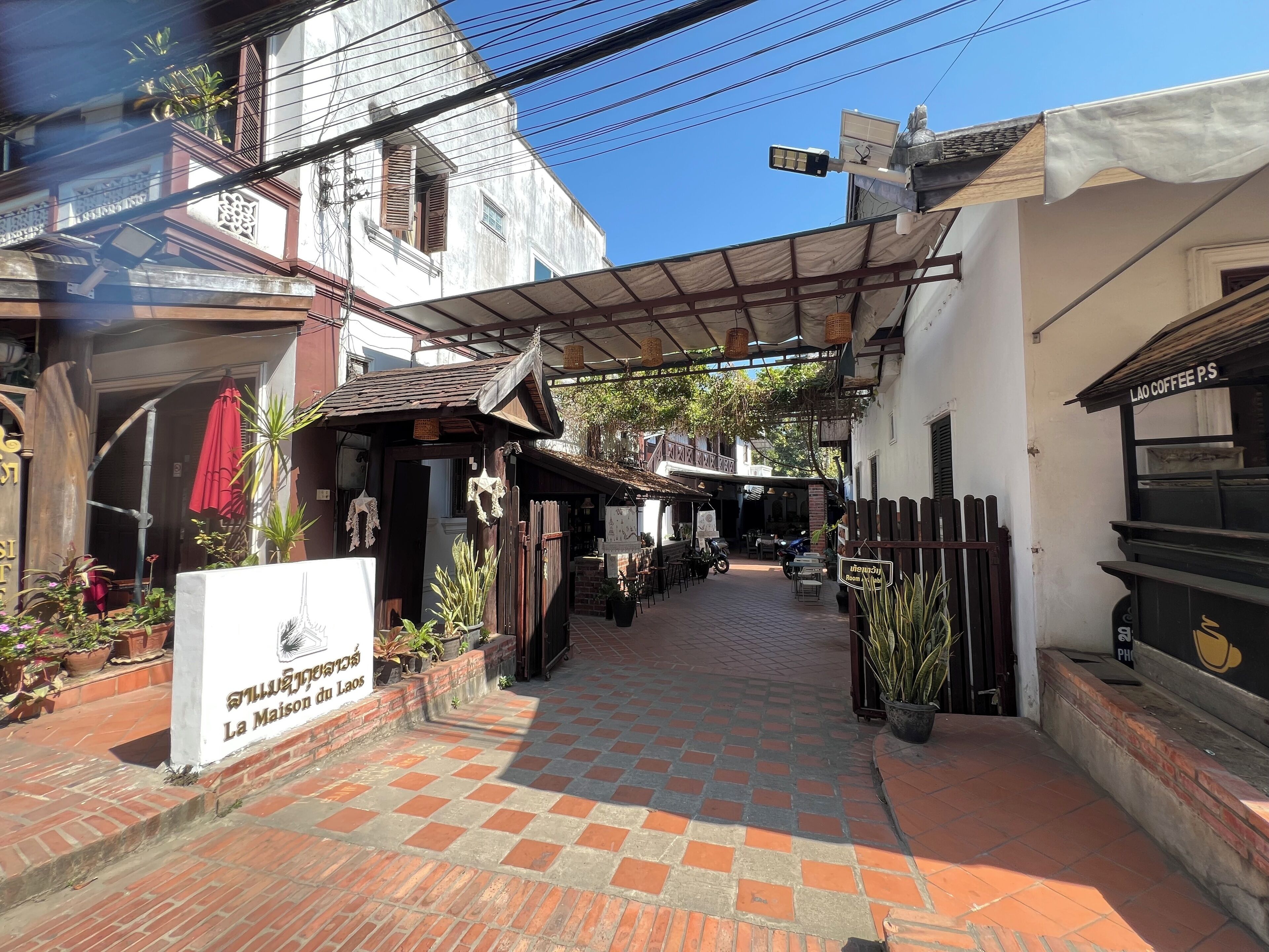 Un estrecho callejón con un sendero adoquinado, un letrero que dice 'La Maison du Laos' y una cafetería llamada 'Lao Coffee P.S.'.