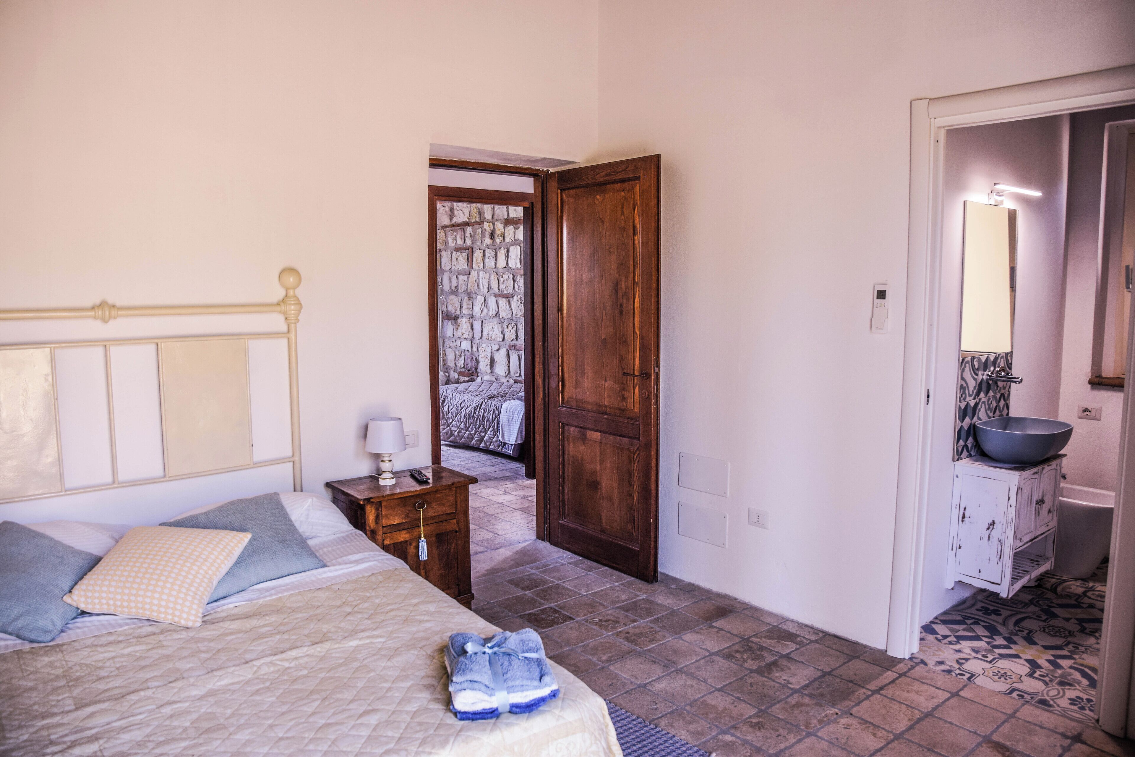 A bedroom with a bed, a nightstand, a wooden door, and a bathroom with a sink and mirror.