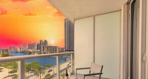 A balcony with a cityscape view at sunset, a chair, and a bottle.