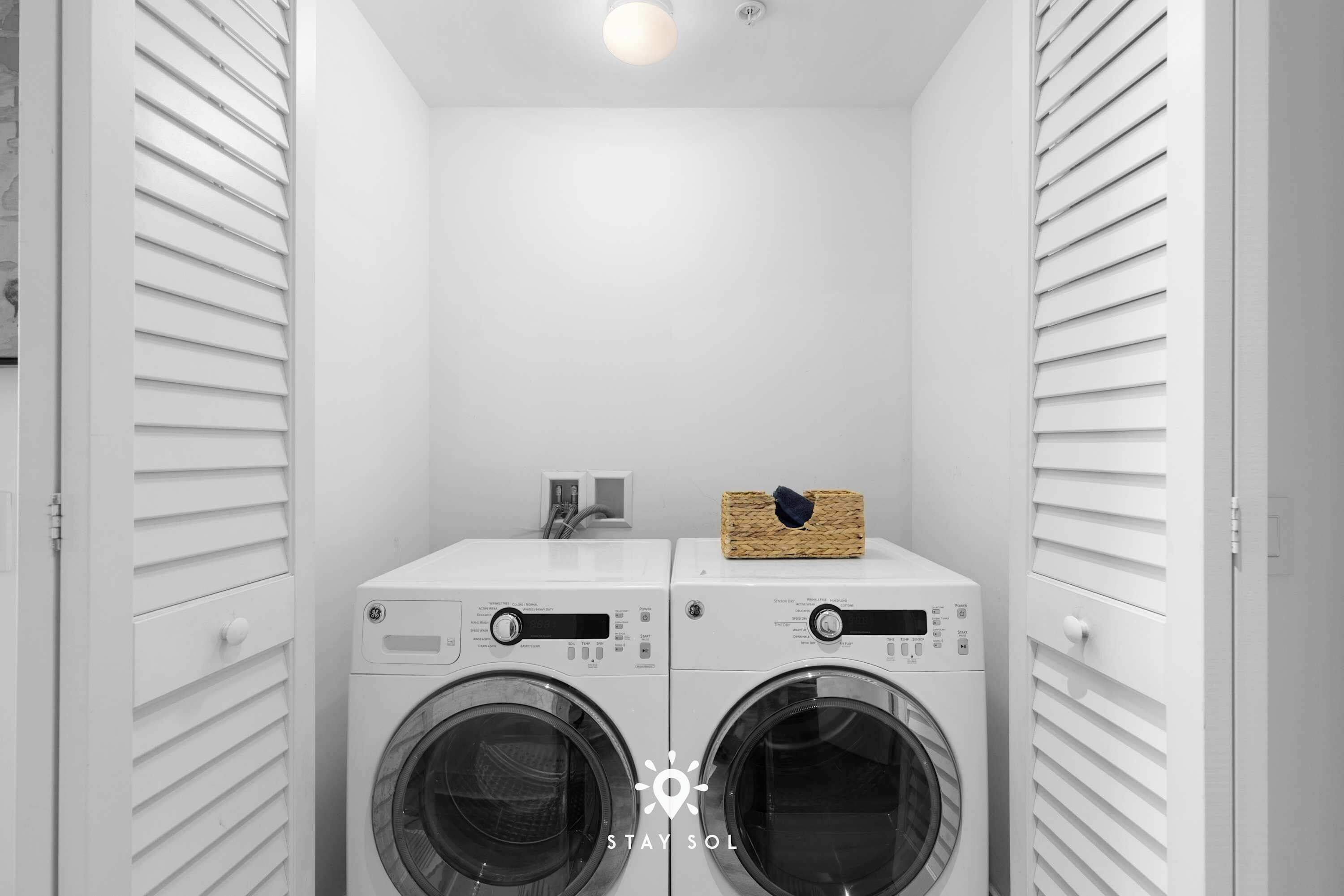 Une buanderie équipée de deux machines à laver à chargement frontal et d’un sèche-linge, avec un panier posé sur le sèche-linge et une prise électrique au mur.
