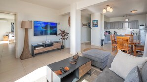 Een moderne woonkamer met een flatscreen televisie, een salontafel en een grijze bank. Er is een eethoek met een houten tafel en stoelen. De keuken is op de achtergrond zichtbaar, met grijze keukenkastjes en een magnetron.