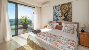 Een slaapkamer met een groot bed, een schilderij van een leeuw, een kamerplant en uitzicht op de oceaan door een schuifdeur.
