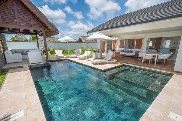 A swimming pool with lounge chairs and umbrellas, a covered seating area, and a kitchen visible through an open door.