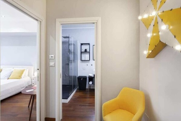 A hotel room with a bed, a small table, a yellow armchair, and a bathroom visible through an open door.
