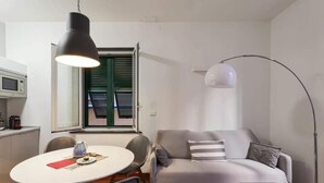 A modern living room with a grey sofa, a round dining table, and a coffee table. There is a large window with blinds, a black pendant light, and a floor lamp. - Libeccio - Style and Comfort at Two Steps from the Cathedral (Genova)