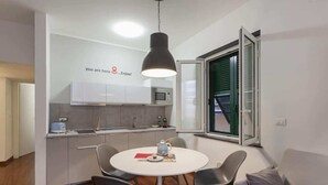 A compact kitchen with modern appliances, a dining area with a round table and chairs, and a large window with blinds. - Libeccio - Style and Comfort at Two Steps from the Cathedral (Genova)