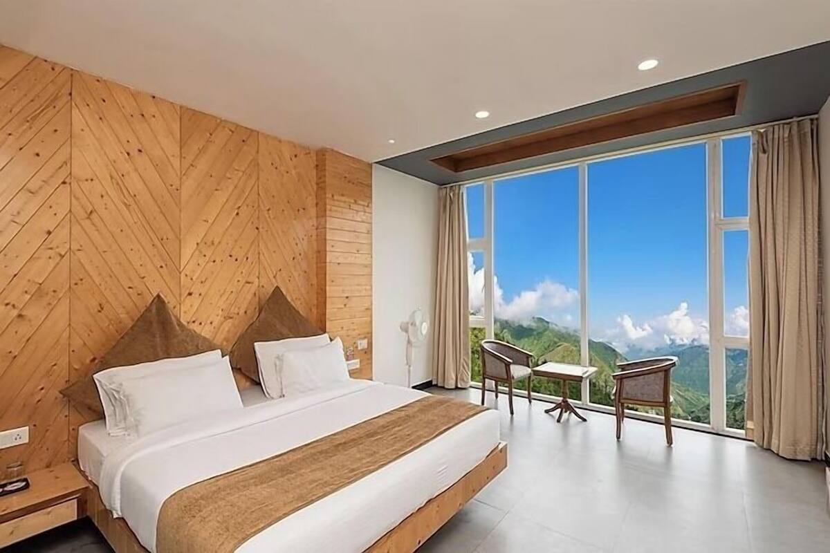 A bedroom with a large bed, wooden wall paneling, a view of mountains through a large window, and a small table with chairs.