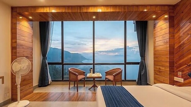 A hotel room with a large window, wooden panel walls, a bed, two chairs, a small table, and a standing fan. - MV Acosta by Lawrence hotels (Mussoorie)