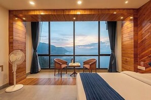 Ett hotellrum med ett stort fönster, väggar beklädda med träpaneler, en säng, två stolar, ett litet bord och en bordsfläkt.