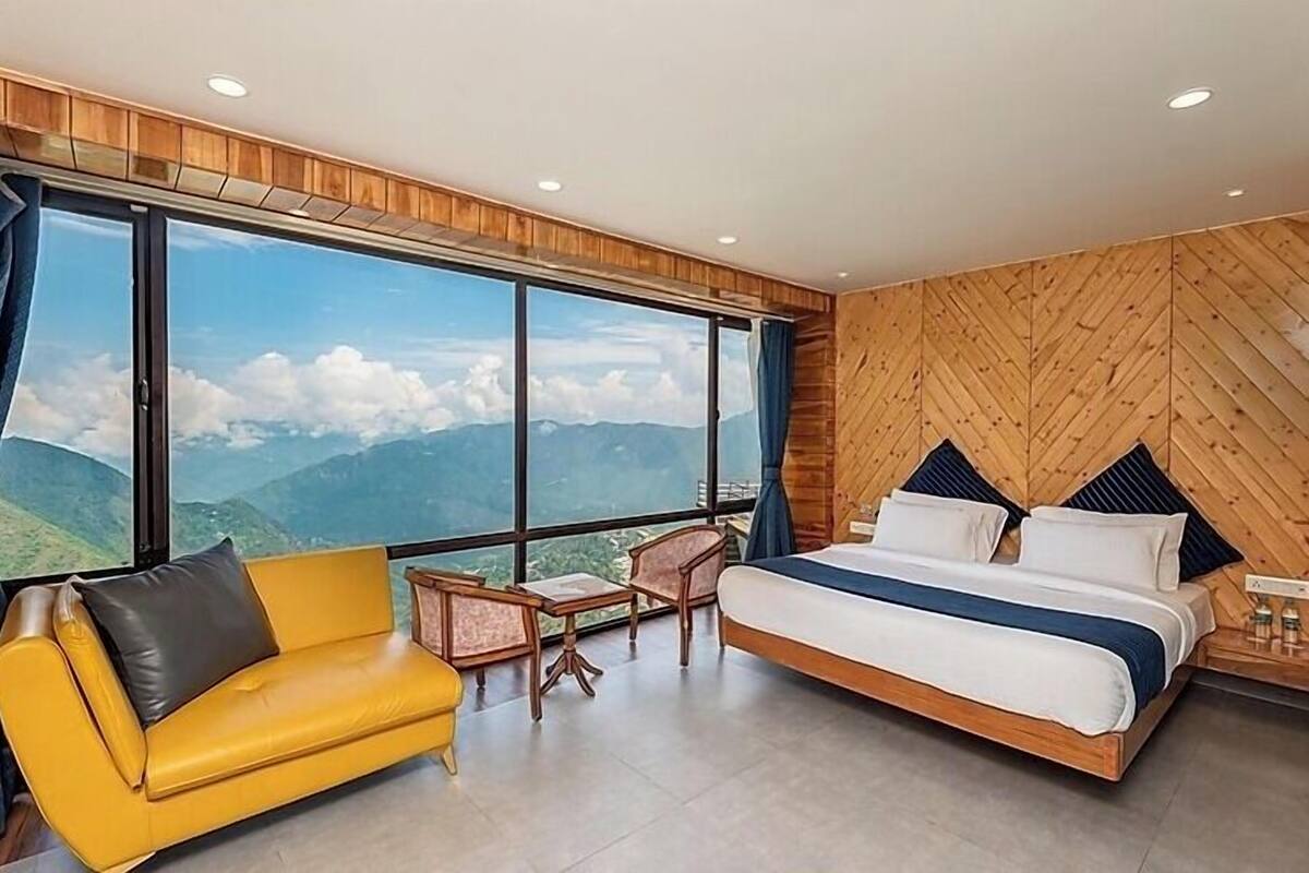 A modern hotel room with a large window offering a mountain view, a bed with white bedding, a yellow sofa, and wooden paneling on the walls.